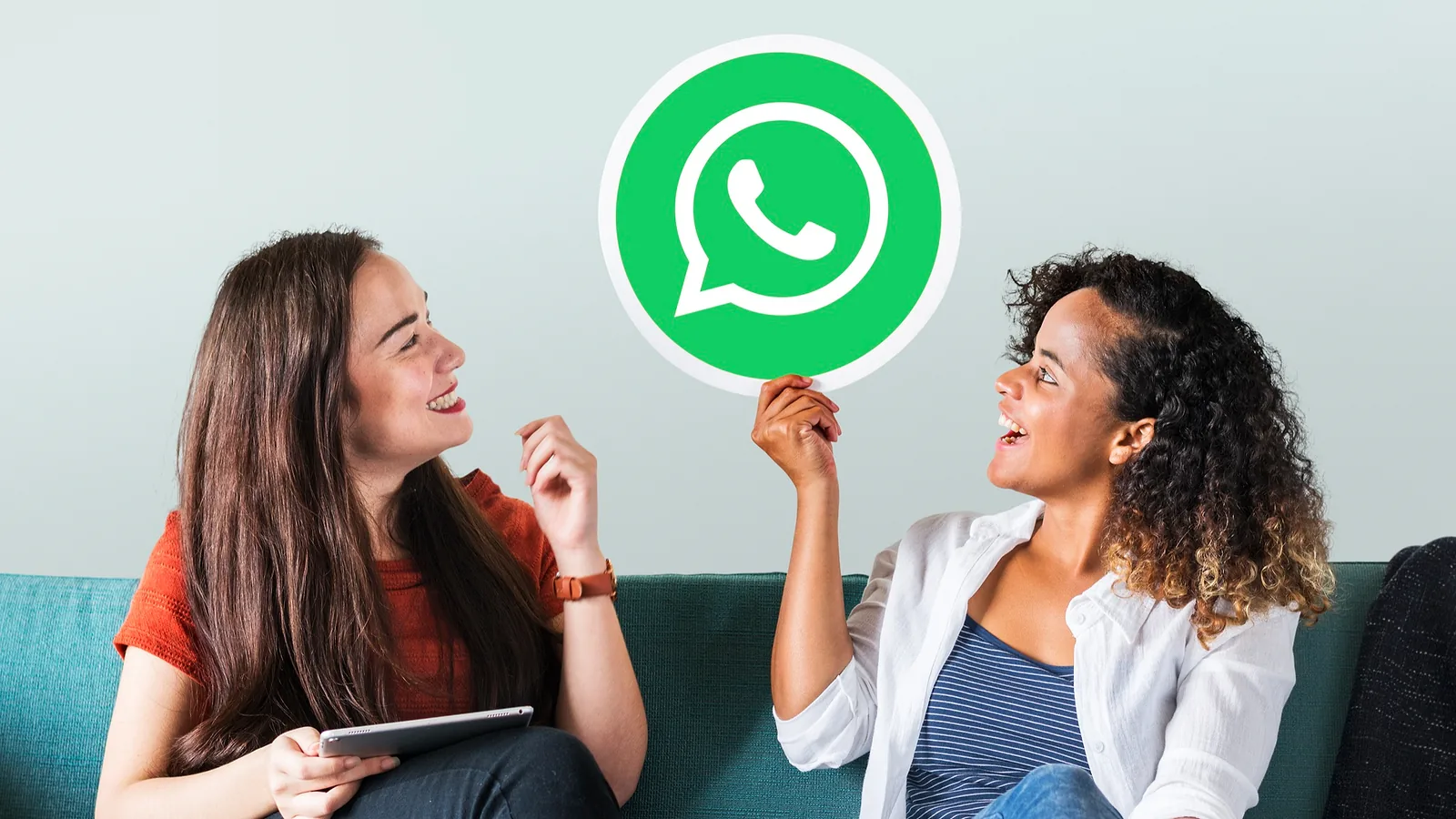 Due amiche sul divano ridono, una tiene in mano un cartello con il logo di WhatsApp